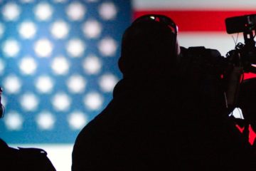 A man with a video camera silhouette; American flag in background.